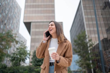 İş kadını telefonda konuşurken gülüyor ve şehir ortamında kahve tutuyor.