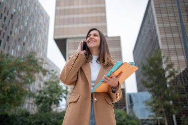 Gülümseyen iş kadını akıllı telefon kullanıyor ve modern ofis binalarının önünde dokümanları tutuyor.