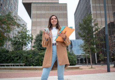 Genç profesyonel kadın elinde klasörlerle gülümsüyor ve modern ofis binalarının dışında akıllı telefon kullanıyor.