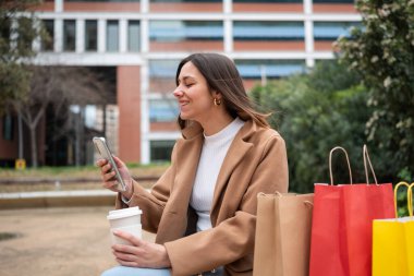 Genç bir kadın alışveriş çılgınlığından sonra akıllı telefonunu kullanırken kahve içmekten hoşlanır.