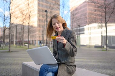 İş kadını bankta oturuyor ve bilgisayarını kredi kartıyla online alışveriş için kullanıyor.