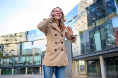 Akıllı telefondan konuşan ve şehir merkezindeki birine el sallayan mutlu iş kadını.