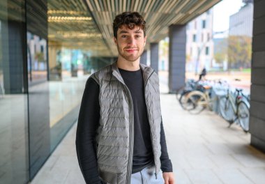 Modern bir şehir ortamında gülümseyen, şık bir yelek giyen kendinden emin bir gencin portresi.