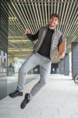 Genç işadamı modern ofis binasının önüne atlıyor, elinde belgeler tutuyor ve başarıyı ve başarıyı ifade ediyor.