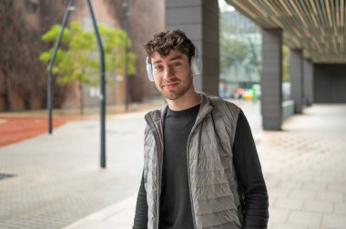 Şık bir öğrenci şehir ortamında yürürken modern kulaklıklar kullanarak müzik dinliyor.