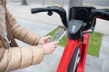 Bir şehirde paylaşılan elektrikli bisikleti açmak için akıllı telefon uygulaması kullanan bir kadın sürdürülebilir ulaşımı teşvik ediyor.