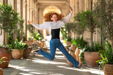 Kaygısız kızıl saçlı kadın sevinçten zıplıyor kulaklıkla müzik dinliyor ve oyun salonunda akıllı telefon tutuyor.