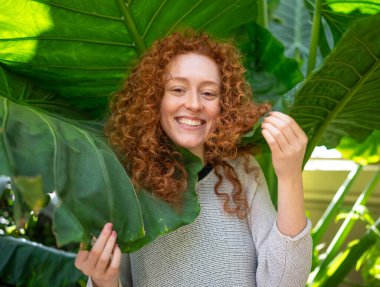 Yeşil yaprakların arasında kıvırcık saçlı neşeli kızıl saçlı genç kadın.