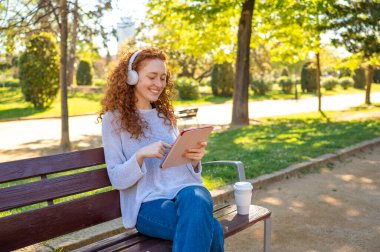 Dijital tablet kullanarak ve müzik dinleyerek gülümseyen kadın, parkta bir bankta oturuyor.