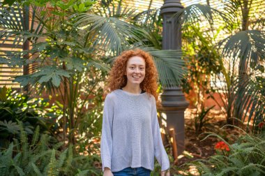 Botanik bahçesindeki mutlu kızıl saçlı kadının portresi, tropikal bitkilerle çevrili.