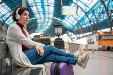 Bir istasyonda trenini beklerken kulaklıkla müzik dinleyen bir turist.