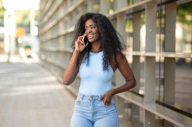 Genç siyahi kadın akıllı telefonuyla konuşurken gülümsüyor, şehirde yürüyor.