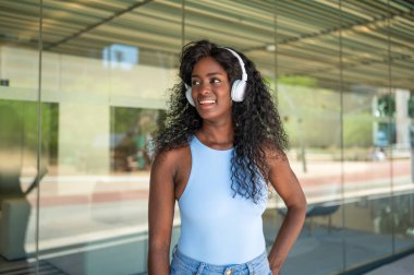 Uzun kıvırcık saçlı, gülümseyen siyah kadın şehir bağlamında kulaklıklı müzik dinliyor.