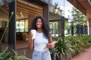 Kendine güvenen genç yönetici modern ofis binasının önünde yürüyor, kahve içiyor ve elinde tablet tutuyor.