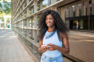 Şehirde modern cam binanın yanında yürürken cep telefonu kullanan genç siyahi bir kadın.
