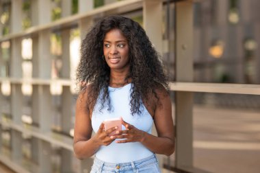 Uzun kıvırcık saçlı, akıllı telefonunu kullanan ve şehirde yürürken gülümseyen genç siyah kadın.