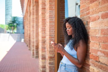 Genç siyahi kadın akıllı telefon kullanıyor. Duvara yaslanıyor.