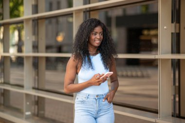 Gülümseyen Afrikalı Amerikalı kadın şehir caddesinde yürürken cep telefonu kullanıyor.
