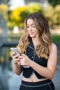 Şehir parkında cep telefonu kullanan gülümseyen genç kadın çevrimiçi bağlantının ve iletişimin tadını çıkarıyor.