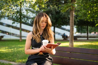 Şehir parkında bankta otururken tablet kullanan ve kahve içen konsantre iş kadını.