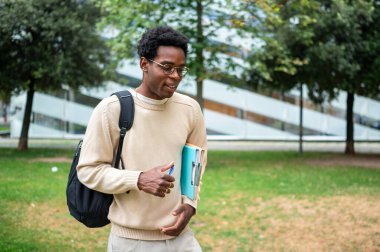 Mutlu Afro-Amerikan üniversite öğrencisi elinde dosyalar ve kalemle kampüs parkında yürüyor.