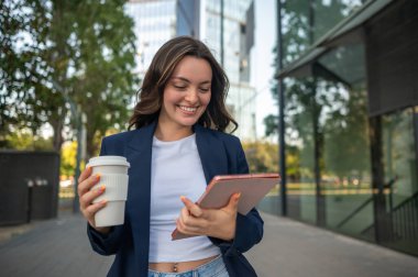Genç yönetici ofis binasının önünde yürüyor, elinde kahve ve tablet kullanıyor.
