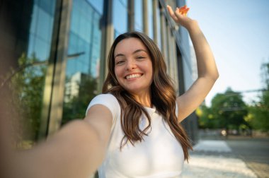 Modern bir ofis binasının önünde kollarını açıp selfie çeken mutlu genç bir kadın.