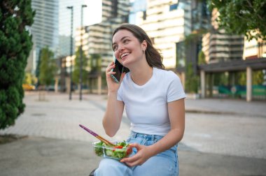 Şehir parkında öğle tatilinin tadını çıkarırken cep telefonu kullanan ve salata yiyen genç bir iş kadını.