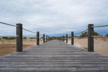 Sahile giden ahşap bir yürüyüş yolunun manzarası bulutlu bir gökyüzünün altındaki kumulları aşıyor.