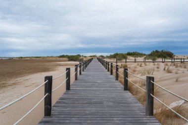 Bulutlu bir gökyüzünün altında kumlu kumsallara doğru uzanan ahşap yürüyüş yolu keşif ve sükuneti davet ediyor.