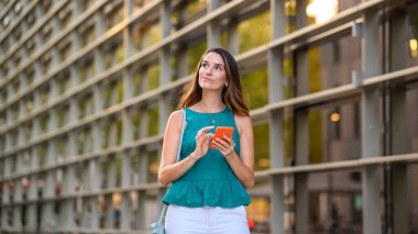 Akıllı telefon kullanan ve modern ofis binasının yakınına bakan dalgın bir iş kadını gelecekteki iş kararlarını düşünüyor.