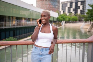 Gülümseyen genç kadın, şehir parkında korkuluklara yaslanırken bir telefon görüşmesi yapıyor.