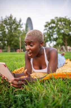 Genç bir kadın çimenlerin üzerinde yatıp tablette şaşırtıcı haberler okuyor. Arka planda da bir bar kulesi var.