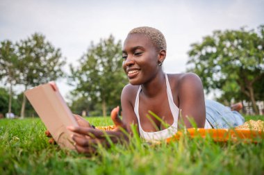 Gülümseyen siyah kadın parkta çimlerin üzerinde uzanırken kitap okumaktan zevk alıyor, doğanın sükunetini kucaklıyor.