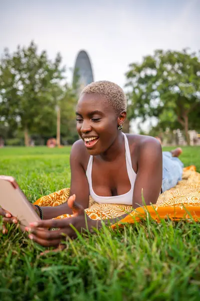Tablet kullanan genç bir kadın, parkta çimlere uzanmış, dışarıda eğleniyor.