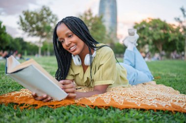 Genç gülümseyen öğrenci parkta çimlerin üzerinde uzanırken kitap okumaktan zevk alıyor.