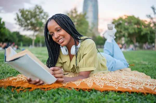 Genç gülümseyen öğrenci parkta çimlerin üzerinde uzanırken kitap okumaktan zevk alıyor.