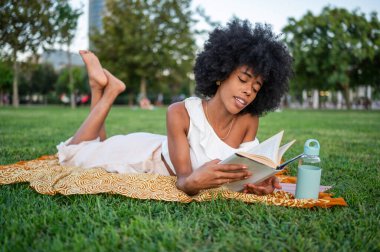 Young woman enjoying reading a book while lying on grass in a park, promoting relaxation and literacy
