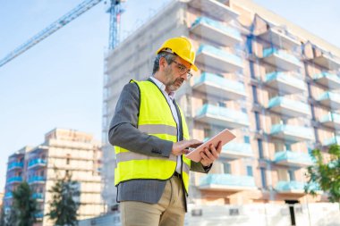 Engineer wearing safety vest and hard hat reviewing construction project plans on a tablet