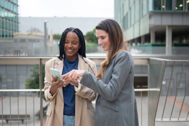 Two businesswomen smiling, collaborating and pointing at a smartphone screen outside corporate buildings