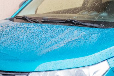 Arabada yağmur damlaları var. Araba parçası yağmur damlalarıyla yakın plan. Yağmur damlalarıyla kaplı mavi bir arabanın kaputu, aynası ve camı. Arabadaki yağmur damlaları çok yakındı.