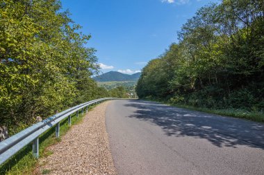 Dağlık arazide asfalt yol. Kayalık dağlar ve güneşli havada sisle kaplı yamaçlar. Otoyolda giden araba, mavi gökyüzünün altında, beyaz kümülüs bulutlarıyla büyük yeşil tepeler arasında uzanıyor.