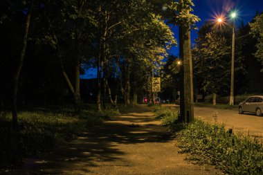 Yazın fenerli gece parkı. Yaz parkının yanındaki yol. Parkın yakınındaki otoyol. Yaz parkının yakınına park edilmiş arabalar. Geceleri fenerlerle aydınlatılmış bir park yolu..