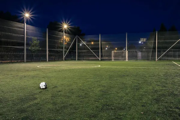 An amateur soccer field with ball illuminated at night. A small football field lit by lanterns in the evening. Green football field illuminated at night. Soccer field in night with spotlight