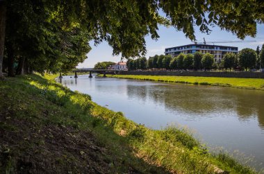 Arka planda bir bina olan nehri geçen bir köprünün manzarası, yemyeşil bitkiler, berrak gökyüzü ve sakin doğal bir çevreye karşı kurulmuş.