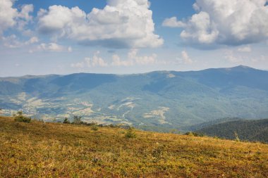 Bereketli bitki örtüsünün, dolambaçlı yolların ve bulutlu mavi gökyüzünün yer aldığı bir dağ manzarası..
