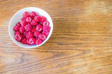 Ahşap bir masaya yerleştirilmiş beyaz bir kasede yüksek açılı taze ahududu fotoğrafı. Sağlıklı yemek temaları için mükemmel.