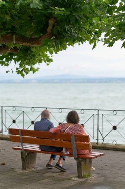    Meersburg, Almanya - 31 Temmuz 2024: Meersburg 'da Constance Gölü kıyısında bir bankta oturan ve manzaranın tadını çıkaran yaşlı bir çift                           