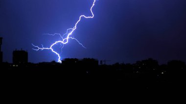 Powerful lightning strike over a dark stormy sky at night over city skyline. Dramatic natural phenomenon with bright blue electric bolts and rain clouds.
