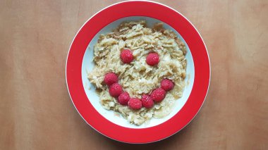 Müsli ve rendelenmiş elmalı çocuk tabağı. Ahududududan yapılmış komik bir yüz. Tahta bir masada..